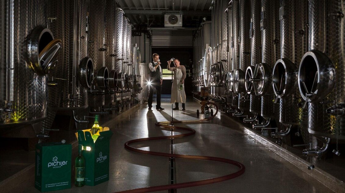 Eine moderne Weinproduktionsanlage mit silbernen Tanks. Zwei Personen stehen inmitten der Tanks und halten ein Gespräch, während eine Schlauchleitung und Produkte im Vordergrund sichtbar sind. | © Bergmann
