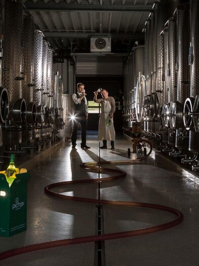 A modern wine production facility with silver tanks. Two people are standing among the tanks engaged in a conversation, while a hose and products are visible in the foreground. | © Bergmann
