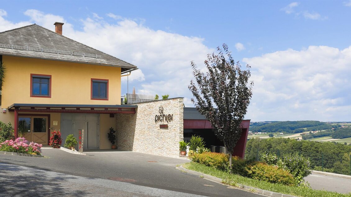 Eine moderne Unterkunft mit gelbem Fassadenanstrich und einem einladenden Eingangsbereich. Im Hintergrund erstrecken sich sanfte Hügel unter einem blauen Himmel. | © Christoph Pohlner