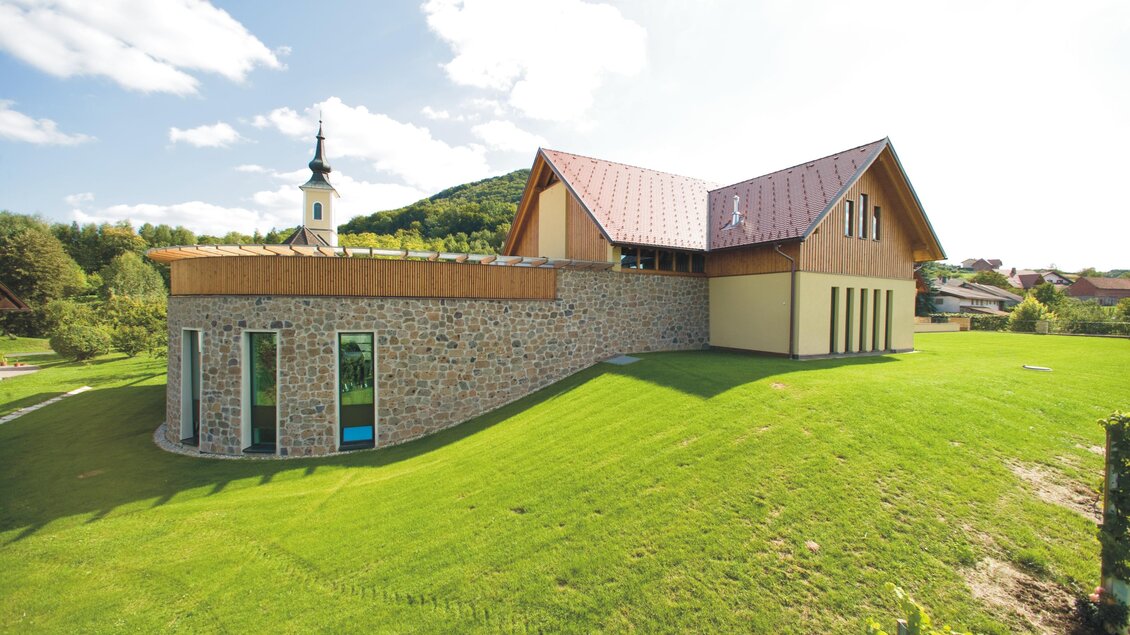 Ein modernes Gebäude mit einem asymmetrischen Dach und Steinmauer. Im Hintergrund sieht man einen Kirchturm und gepflegte Grünflächen. | © Weinhof Platzer