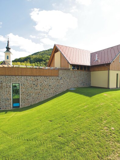A modern building with an asymmetrical roof and stone wall. In the background, you can see a church tower and well-maintained green spaces. | © Weinhof Platzer