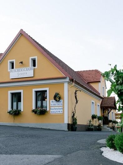 A charming guesthouse with a yellow facade and flowering windows. Next to the building stands an informative sign. | © Buschenschank Sammer