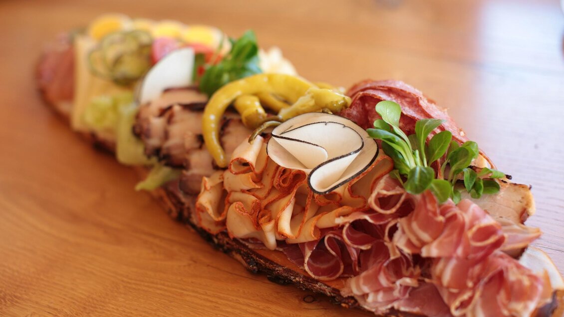 Ein appetitlicher Wurst- und Käseteller auf einem Holzbrett. Verschiedene Aufschnittsorten sind mit Gemüse und Senf dekoriert. | © Familie Ramminger
