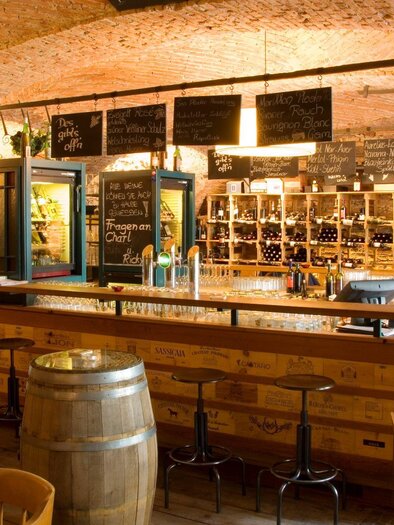 Eine gemütliche Weinbar mit einem langen Bartresen und Regalen voller Weine. Die Wände sind aus Ziegelstein, was eine warme Atmosphäre schafft. | © Klarmann