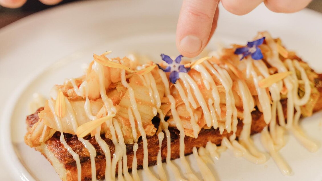 Eine kunstvoll angerichtete Speise auf einem Teller. Ein Koch verziert das Gericht mit einer Soße und essbaren Blüten. | © Weinrefugium Brolli