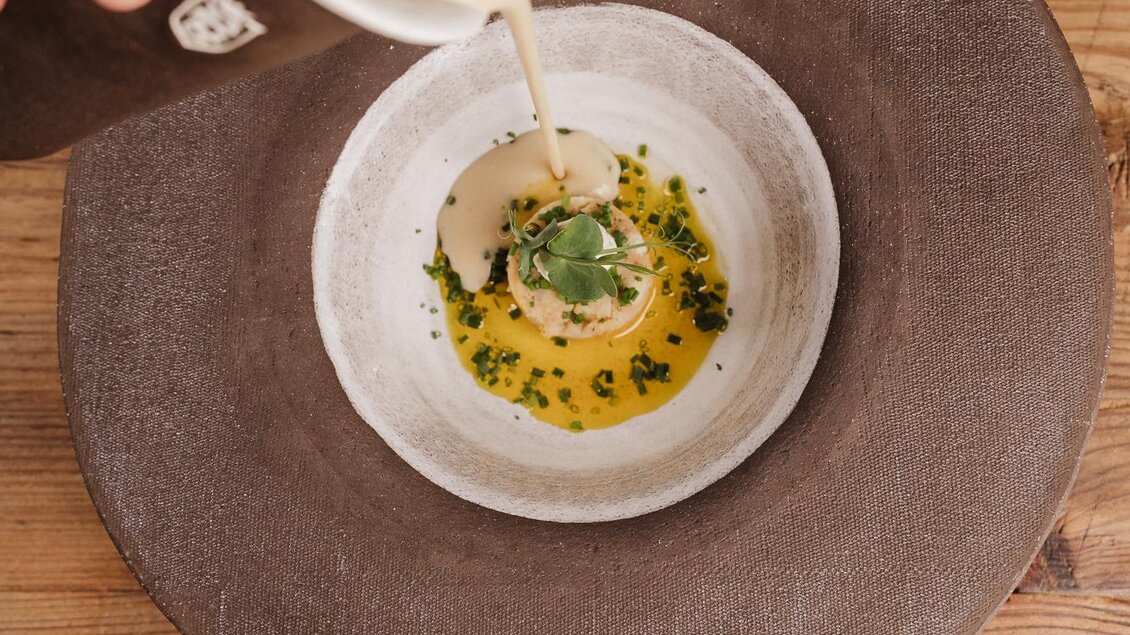 Eine elegante Schüssel mit einem Gericht, das mit einer Flüssigkeit übergossen wird. Der Teller ist auf einem rustikalen Holztisch platziert. | © Weinrefugium Brolli