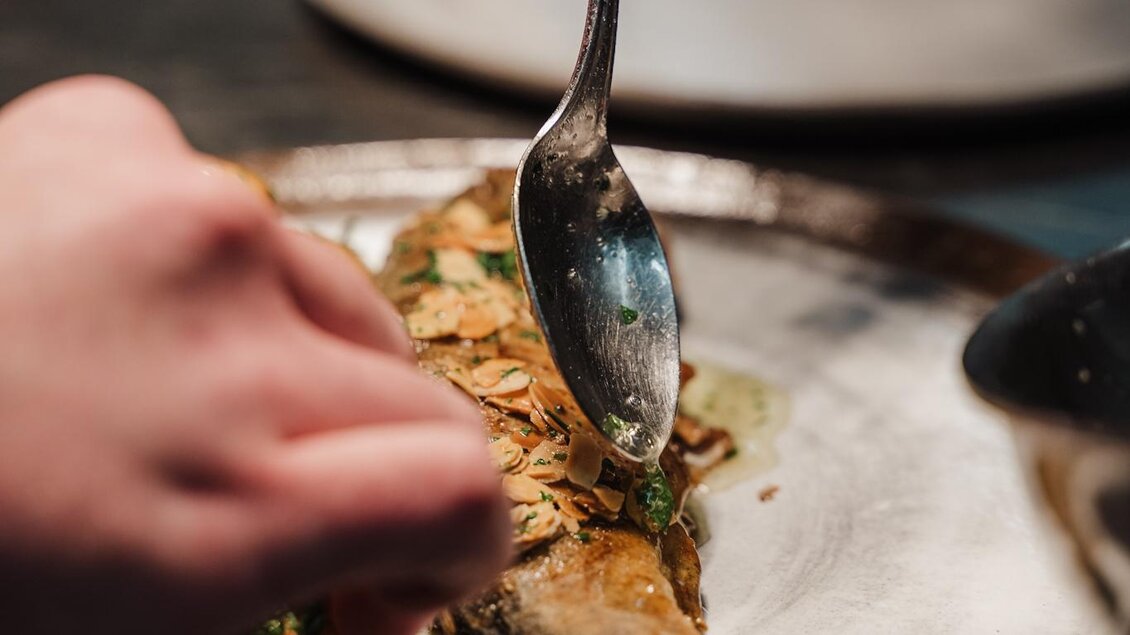 Eine Hand garnisiert einen gegrillten Fisch auf einem Teller. Der Tisch ist dunkel und die Atmosphäre wirkt elegant. | © Weinrefugium Brolli