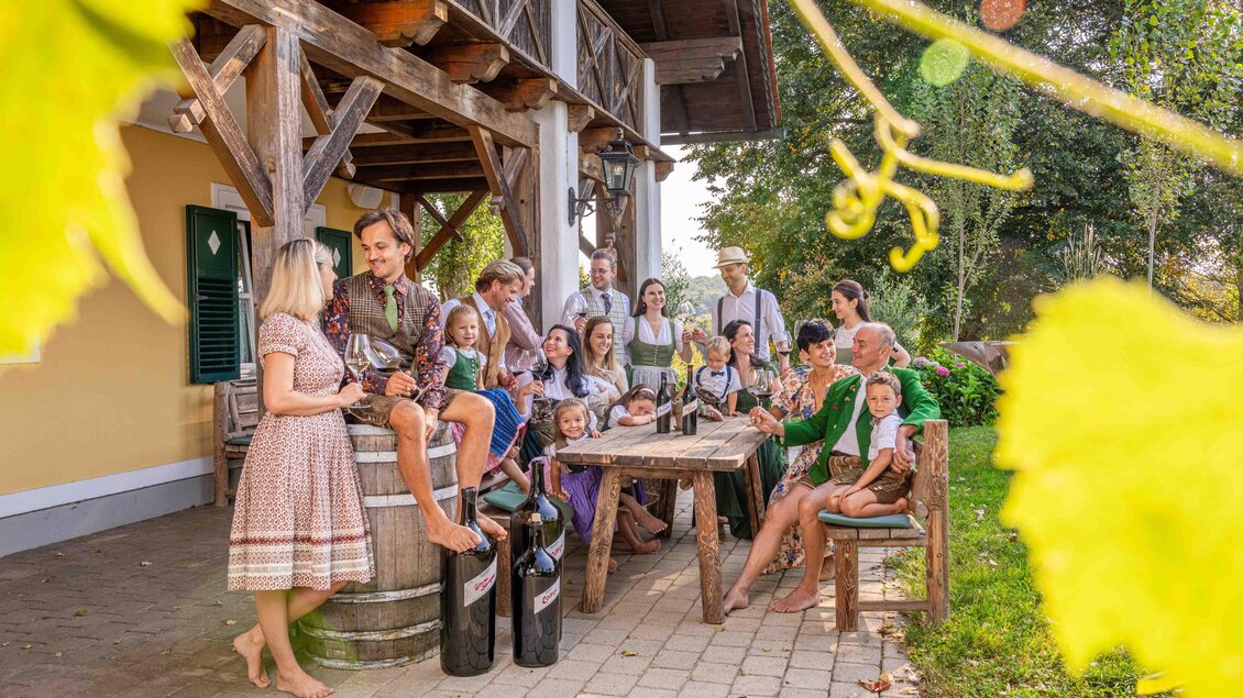 Eine Gruppe von Menschen in traditioneller Kleidung sitzt an einem Tisch im Freien. Im Hintergrund sind ein Holzgebäude und Weinreben zu sehen. | © Weinschloss Thaller