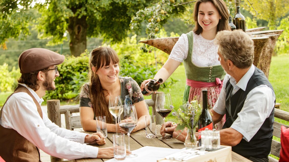 Eine gesellige Runde am Tisch im Freien. Eine Bedienung schenkt Wein an Gäste aus, die in traditioneller Kleidung sitzen.
