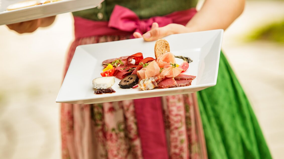 Ein Teller mit verschiedenen Köstlichkeiten, darunter Aufschnitt und frisches Gemüse. Die Person trägt ein traditionelles bayerisches Outfit.