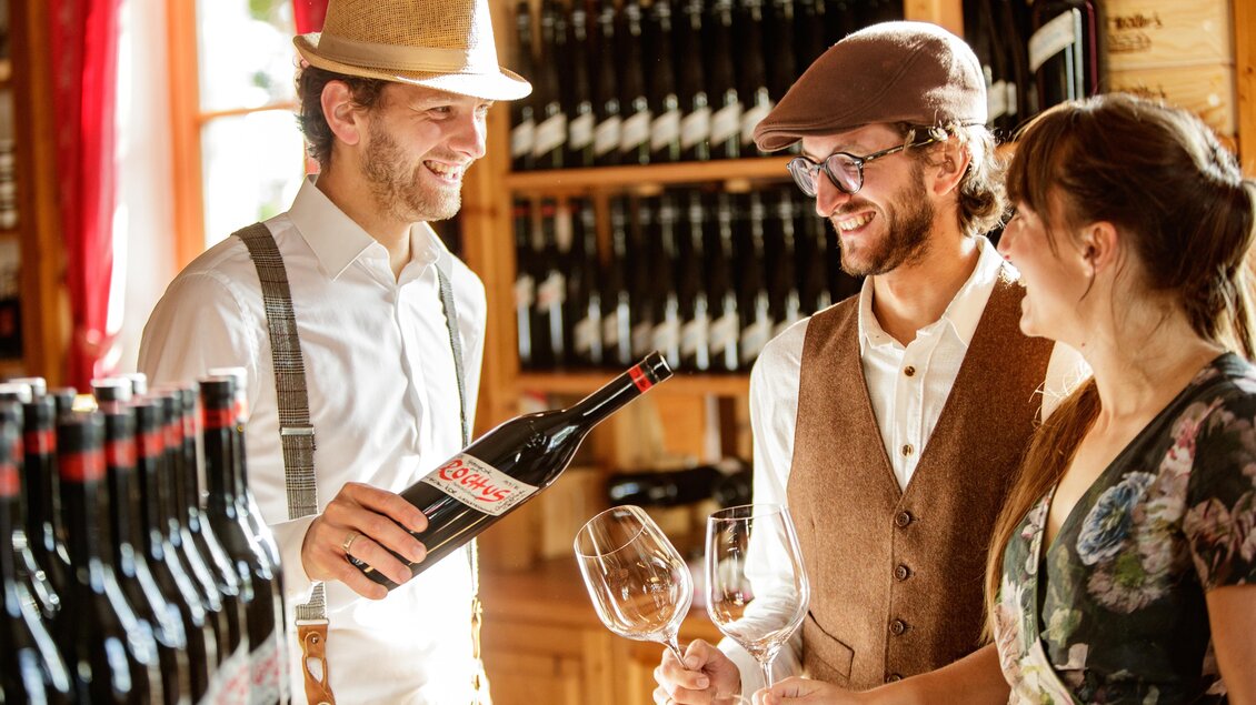 Eine stilvolle Weinverkostung in einem gemütlichen Weingut. Drei Personen lächeln, während sie Weingläser halten und eine Flasche Wein betrachten.