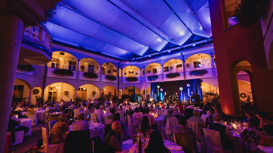 Eine festliche Veranstaltung in einem gut beleuchteten Saal mit vielen Gästen. Die Atmosphäre ist lebhaft, mit Tischen und Stühlen, die liebevoll gedeckt sind. | © Weinschloss Thaller