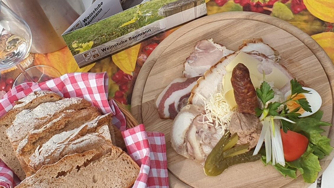 Ein rustikales Brett mit herzhaftem Essen, darunter verschiedene Fleischsorten, Sauerkraut und frisches Gemüse. Neben dem Brett liegt ein Laib Brot mit einer karierten Serviette. | © Weinstube Baumann