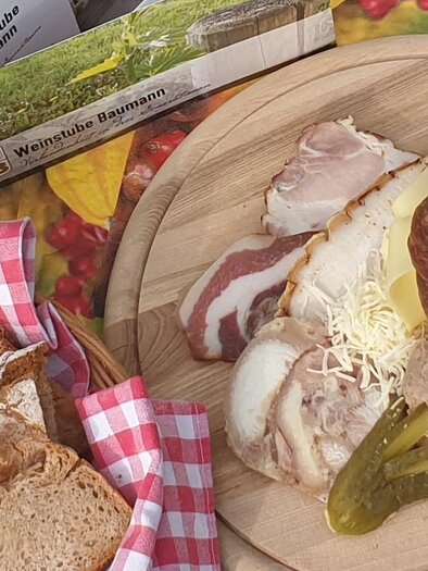 A rustic board with hearty food, including various types of meat, sauerkraut, and fresh vegetables. Next to the board is a loaf of bread with a checked napkin. | © Weinstube Baumann