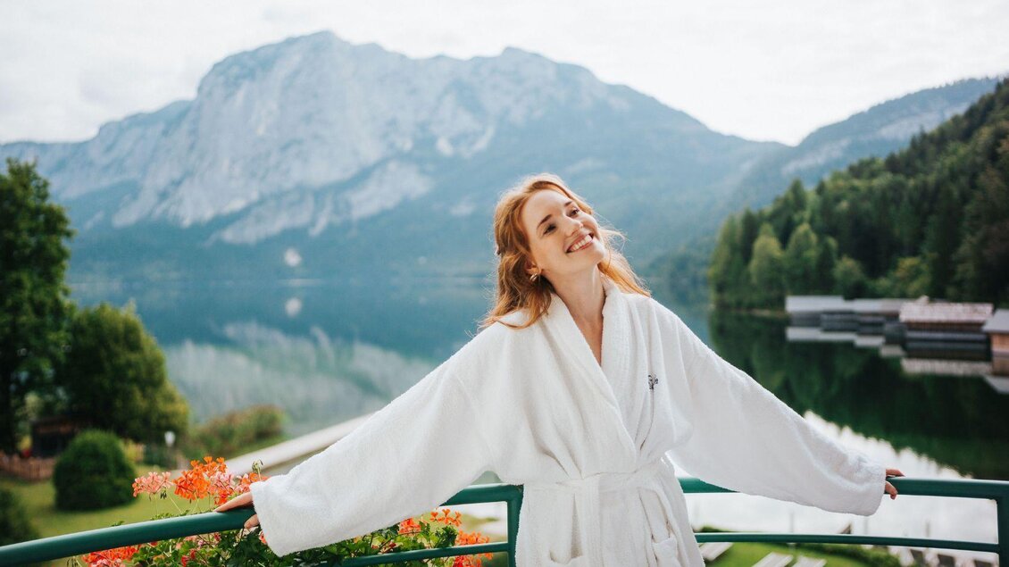 Eine fröhliche Frau in einem weißen Bademantel steht auf einem Balkon mit Blick auf einen malerischen See und beeindruckende Berge. Die Szene strahlt Ruhe und Entspannung aus. | © Karl Steinegger