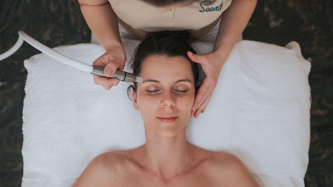 Eine Frau liegt entspannt auf einer Liege, während eine Behandlerin eine Gesichtsbehandlung durchführt. Das Ambiente ist beruhigend und gepflegt. | © Karl Steinegger