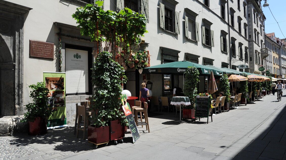 Eine charmante Straße mit gemütlichen Terrassen und grünen Pflanzen. Die Gebäude sind im traditionellen Stil gehalten und laden zum Verweilen ein. | © Welscher Stub'n
