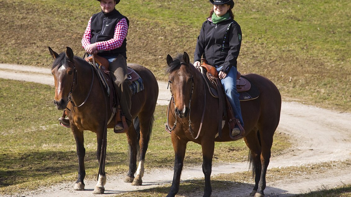Zwei Reiter auf braunen Pferden auf einem Pfad in der Natur. Die Reiter tragen passende Kleidung und Cowboyhüte. | © Lasingerhof