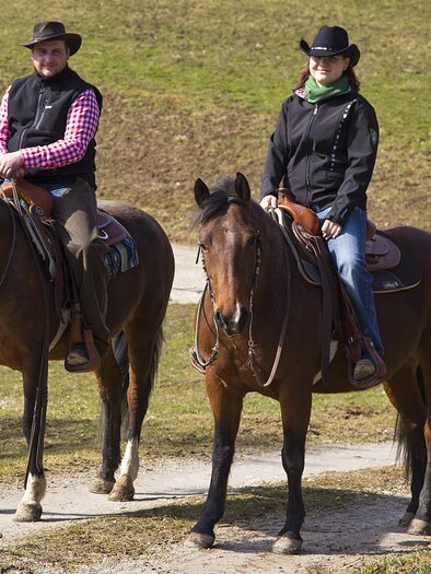 Two riders on brown horses on a path in nature. The riders are wearing matching outfits and cowboy hats. | © Lasingerhof