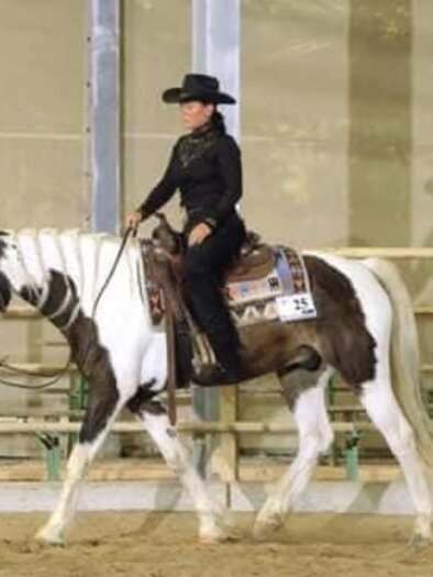 Eine Reiterin in schwarzer Kleidung sitzt auf einem zweifarbigen Pferd in einer Halle. Im Hintergrund stehen Zuschauer, die das Geschehen beobachten. | © Tina Edenhofer