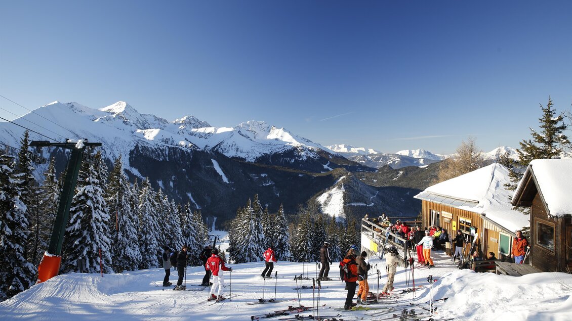 Eine malerische Skilandschaft mit verschneiten Bergen und Bäumen. Skiende Personen genießen den klaren Himmel und die Sonne. | © Herbert Raffalt