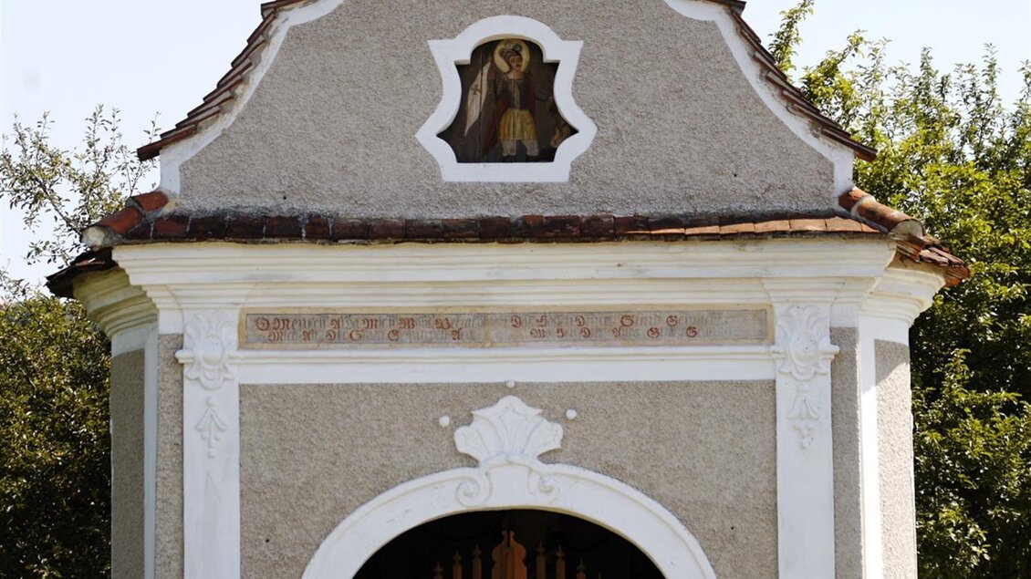 Ein kleiner, historischer Bau mit einer kunstvollen Fassade und einem auffälligen Dachkreuz. Vor der Tür stehen blühende Pflanzen in Töpfen. | © Lederer Josef