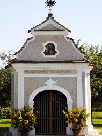 Ein kleiner, historischer Bau mit einer kunstvollen Fassade und einem auffälligen Dachkreuz. Vor der Tür stehen blühende Pflanzen in Töpfen. | © Lederer Josef
