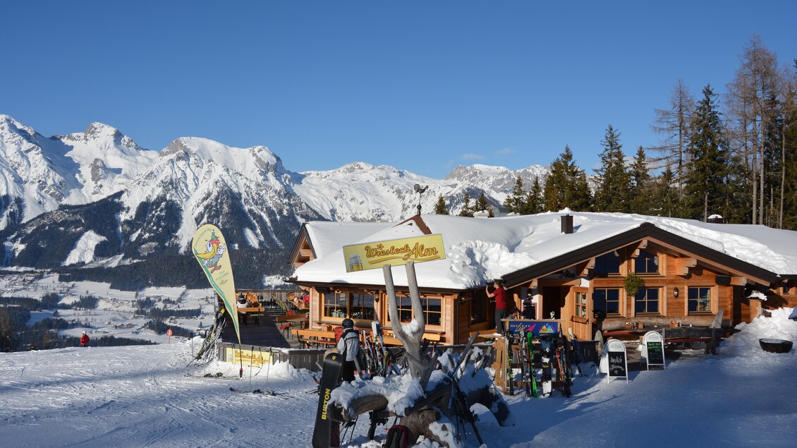 Eine gemütliche Berghütte im Schnee, umgeben von beeindruckenden Bergen und einem blauen Himmel. Skifahrer genießen die winterliche Landschaft. | © eigen