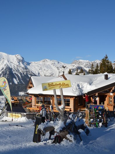 Eine gemütliche Berghütte im Schnee, umgeben von beeindruckenden Bergen und einem blauen Himmel. Skifahrer genießen die winterliche Landschaft. | © eigen