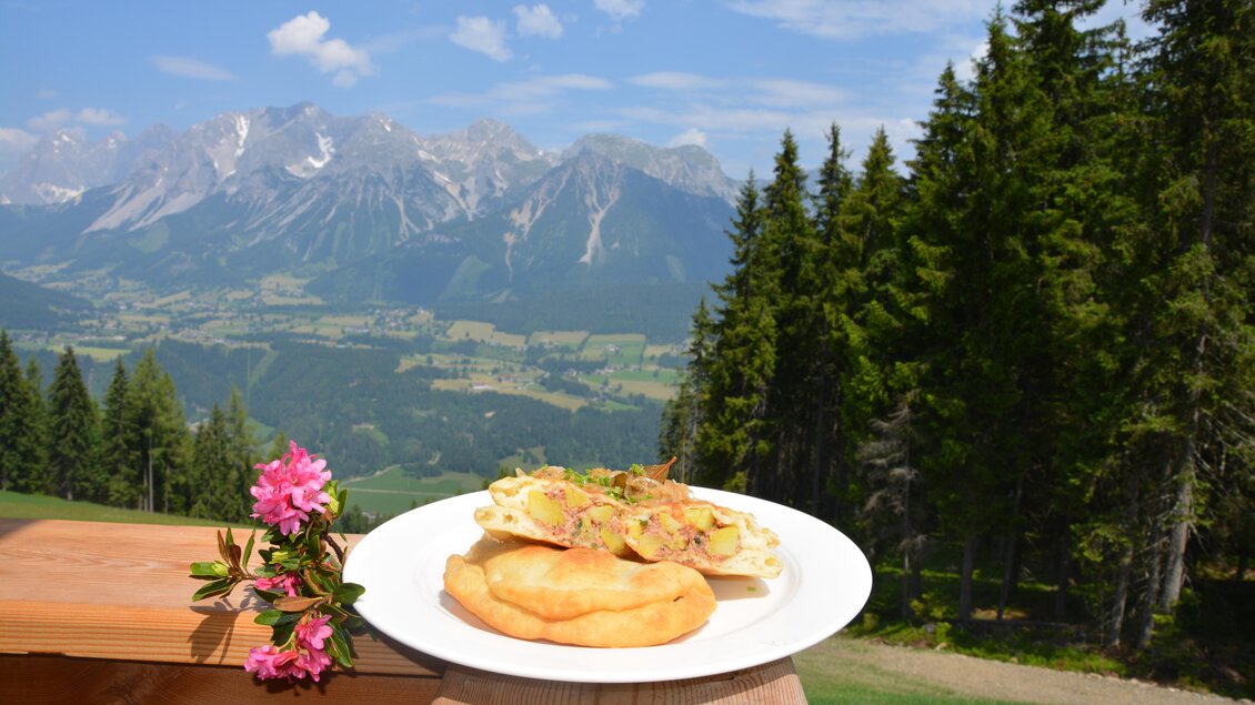 Ein Teller mit frischem Essen steht auf einem Holztisch, umgeben von einer wunderschönen Berglandschaft. Im Hintergrund erkennt man hohe Berge und grüne Wälder unter einem blauen Himmel. | © eigen