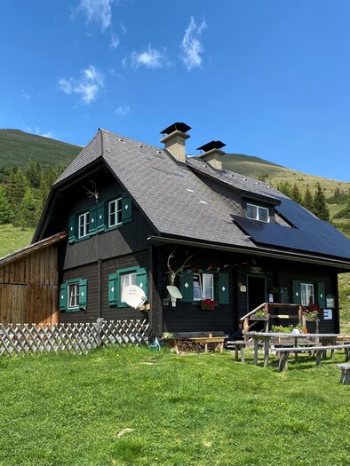 Ein gemütliches schwarzes Haus in der Natur mit grünen Fensterläden. Umgeben von Wiesen und Bergen unter einem blauen Himmel. | © Erlebnisregion Murtal