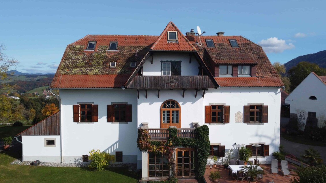 Ein großes, traditionelles Haus mit einem roten Ziegeldach und weißen Wänden. Der Garten ist grün und es gibt einige Pflanzen um das Gebäude herum. | © Jürgen Laban