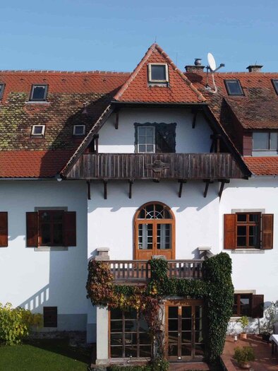 Ein großes, traditionelles Haus mit einem roten Ziegeldach und weißen Wänden. Der Garten ist grün und es gibt einige Pflanzen um das Gebäude herum. | © Jürgen Laban