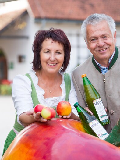 Ein fröhliches Paar steht neben einem großen, bunten Obst-Motiv. Sie halten Äpfel und Flaschen in der Hand und lächeln freundlich. | © Wilhelm Spezialitäten