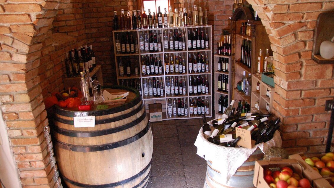 Ein gemütlicher Weinkeller mit Regalen voller Flaschen und Holzfässern. Auf einem Tisch liegen frisches Obst und einige Flaschen Wein. | © Wilhelm Spezialitäten