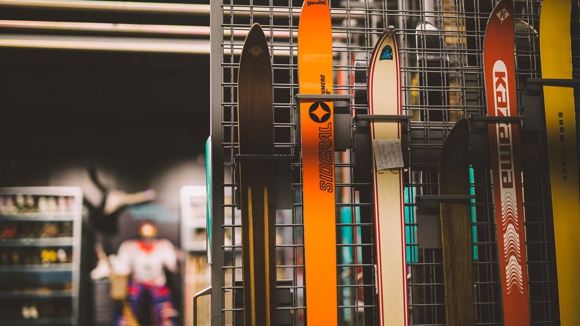 Skier stehen in einem Geschäft an einem Ständer. Im Hintergrund sind weitere Sportartikel sichtbar. | © WinterSportMuseum