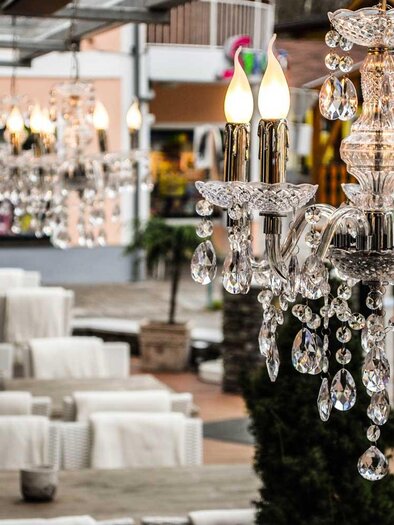 Ein eleganter Kronleuchter aus Kristall hängt über einem stilvollen Außenbereich. Im Hintergrund sind gemütliche Sitzgelegenheiten und grüne Pflanzen zu sehen. | © Wintergarten