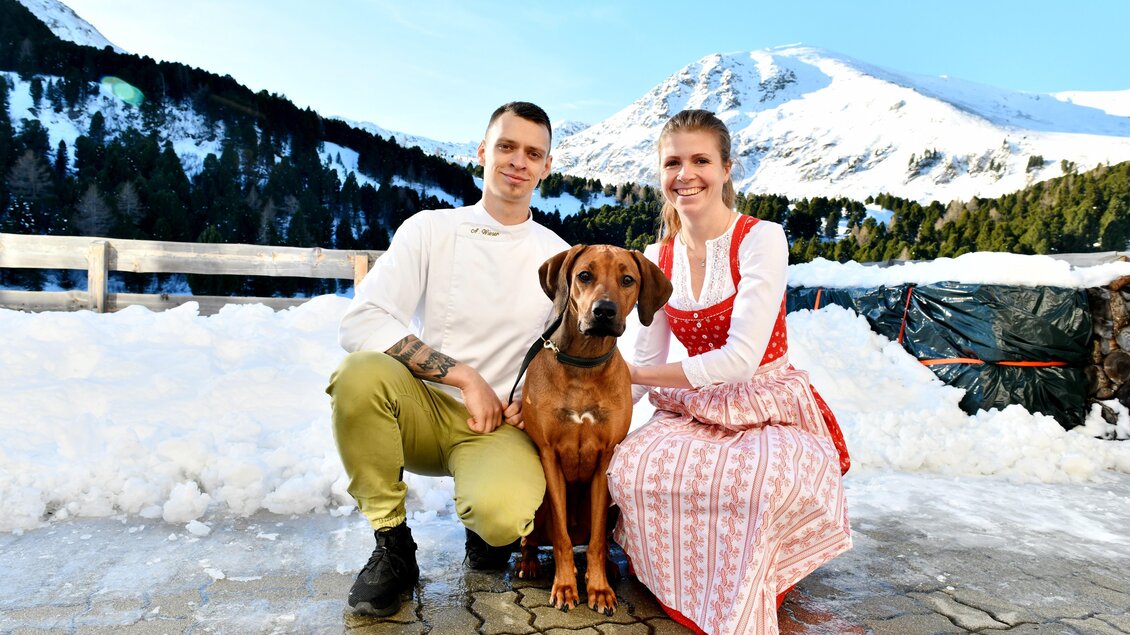 Ein Paar in traditioneller Kleidung sitzt im Schnee mit einem Hund. Im Hintergrund sind Berge und ein blauer Himmel zu sehen. | © Andreas Wieser