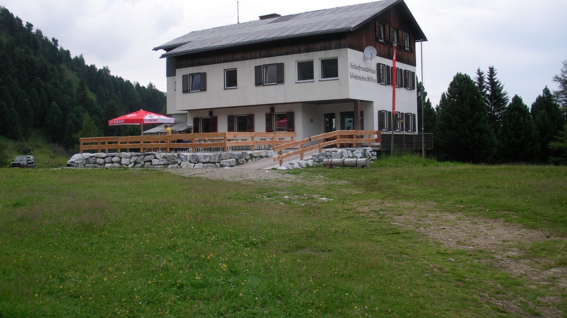 Ein Gebäude in der Natur, umgeben von Wiesen und Bäumen. Es hat eine Terrasse mit Sonnenschirmen und einen rustikalen Charme. | © Andreas Wieser