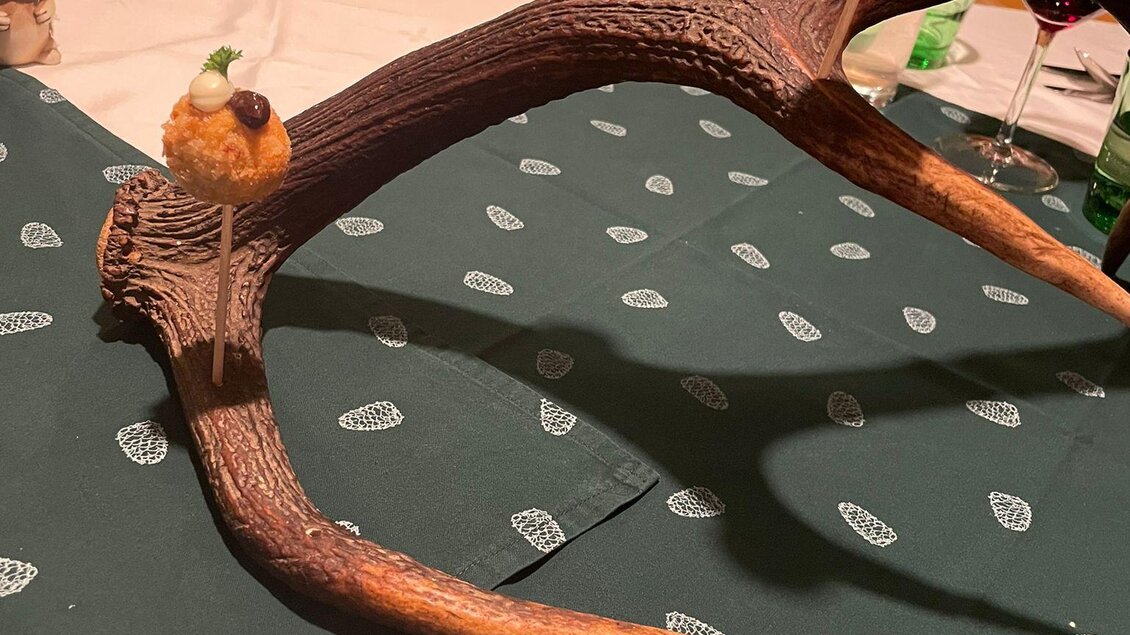 Ein dekoratives Geweih auf einem Tisch, darauf sitzen kleine Köstlichkeiten. Der Tisch ist mit einem grünen Tuch und Stühlen im Hintergrund gedeckt. | © Andreas Wieser