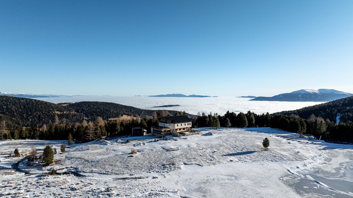 Eine verschneite Landschaft mit einem Gebäude und einer hohen Aussicht auf eine Wolkendecke. Der Himmel ist klar und blau, während die Berge im Hintergrund sichtbar sind. | © Tom Lamm
