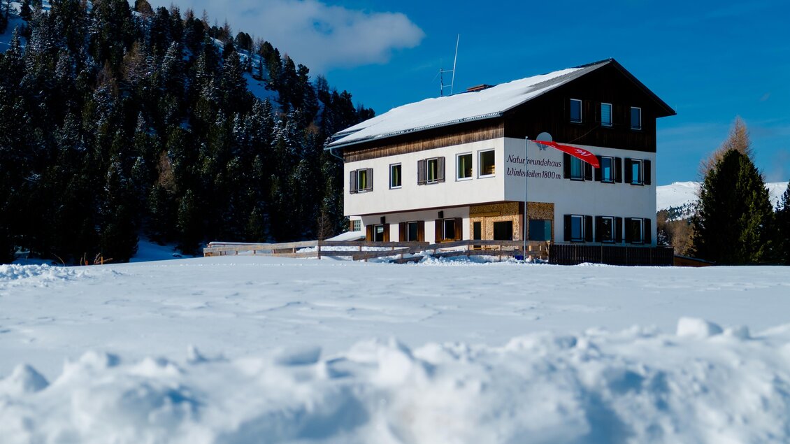 Ein einladendes Gebäude im Schnee, umgeben von grünen Hügeln und bewaldeten Bergen. Der Himmel ist klar und blau, was eine ruhige winterliche Atmosphäre schafft. | © Andreas Wieser