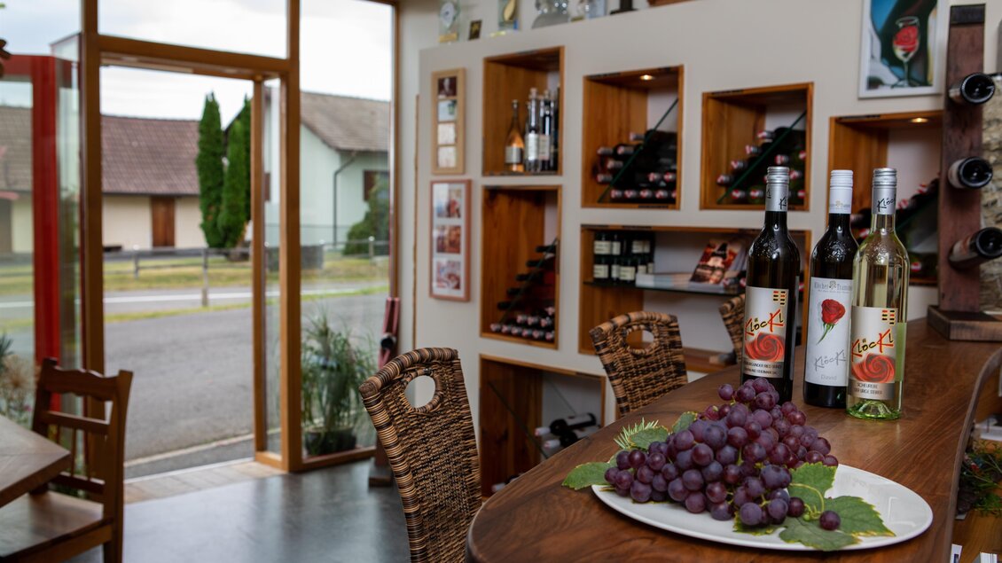 Ein einladender Weinladen mit Regalen voller Weinflaschen. Auf dem Tisch liegt ein Teller mit frischen Trauben. | © Winzerhof Klöckl
