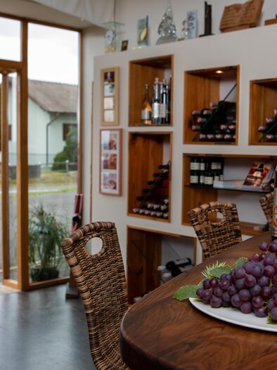 An inviting wine shop with shelves full of wine bottles. On the table lies a plate of fresh grapes. | © Winzerhof Klöckl
