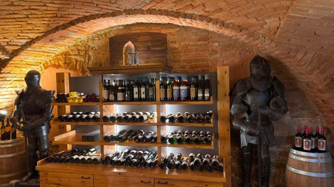 Ein gemütlicher Weinkeller mit einer Holzregal, das mit vielen Weinflaschen gefüllt ist. Auf beiden Seiten stehen Rüstungen aus der Vergangenheit. | © © Linshalm Fotografie
