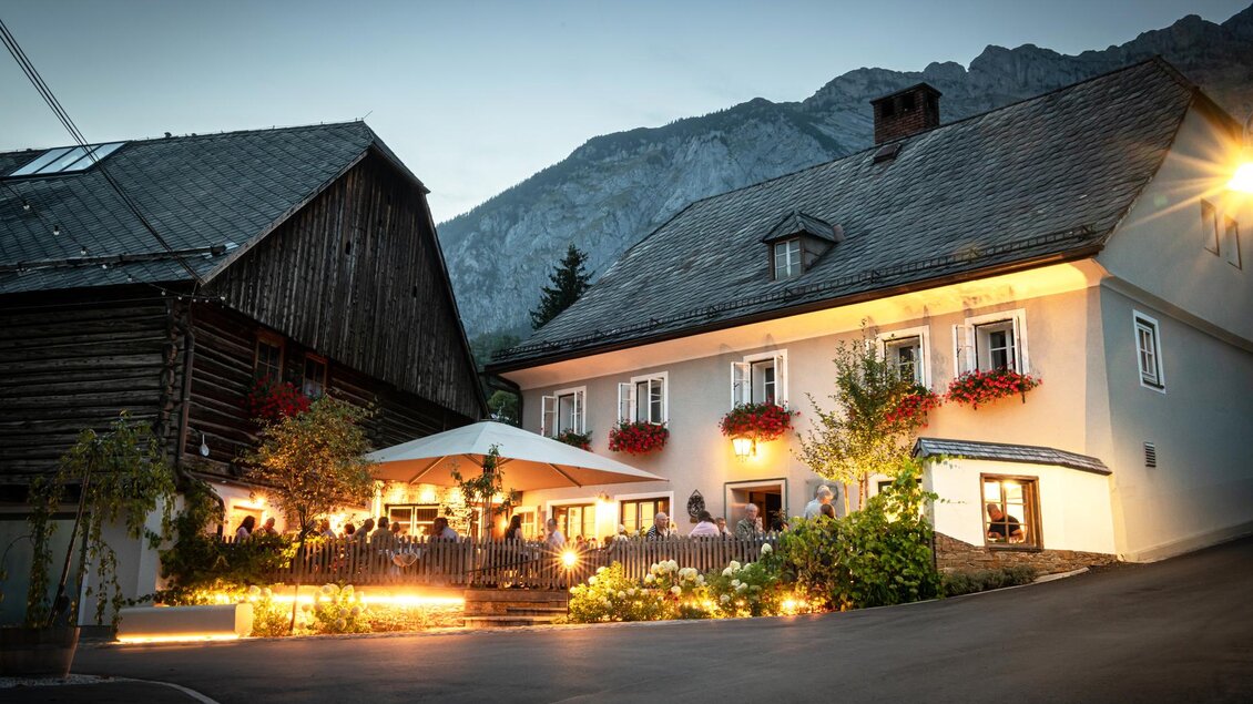 Ein einladendes Gasthaus am Abend mit blühenden Fenstern und sanfter Beleuchtung. Im Hintergrund sind die Berge zu sehen, die eine malerische Kulisse bieten. | © Christoph Huber