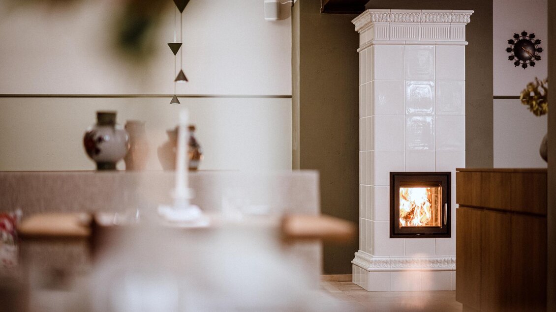 Ein gemütlicher Innenraum mit einem weißen Kachelofen. Im Hintergrund sind dekorative Elemente und ein schöner Tisch zu sehen. | © Christoph Huber