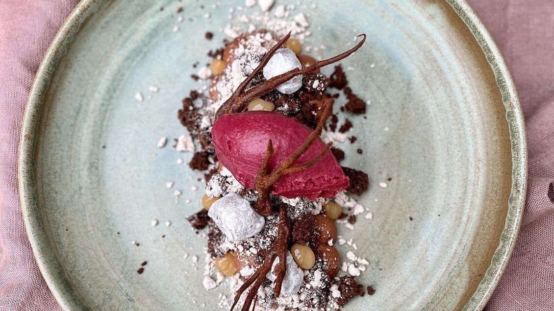 Ein kunstvoll angerichtetes Dessert auf einem grünen Teller. Es enthält eine rosa Eisschicht, Stücke von Schokolade und knusprige Elemente. | © Lisa Ell-Mayer