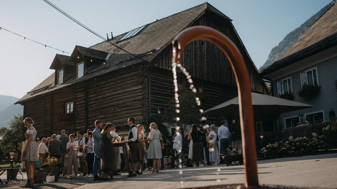 Ein schönes Fest im Freien mit vielen Gästen vor einem rustikalen Holzgebäude. Eine Wasserstation sprudelt, während die Sonne scheinen. | © Marta Urbanelli