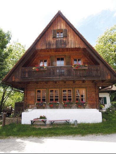 A traditional wooden house with a pointed roof and many windows. It is located in a green environment with trees and a small garden. | © Jagawirt